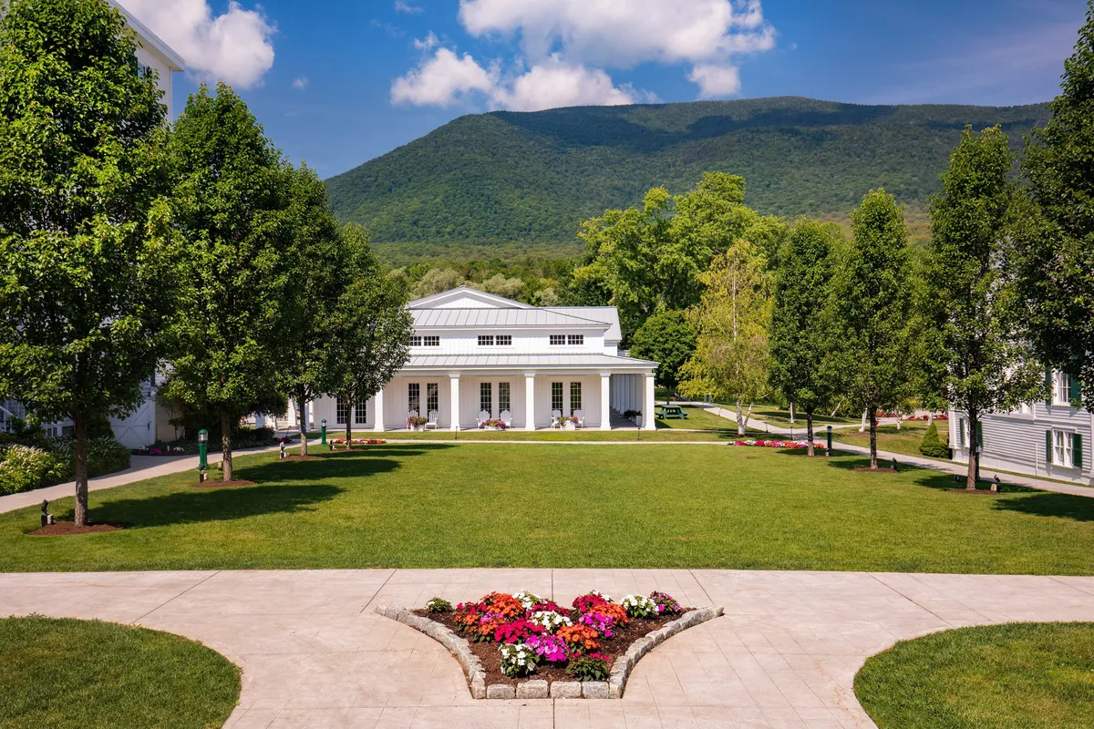 The image showcases a manicured lawn bordered by vibrant flower beds, set against a backdrop of majestic mountains and colonial-style architecture. The space is ideal for outdoor gatherings and quiet strolls, emphasizing the resort’s connection to nature and historic grandeur.