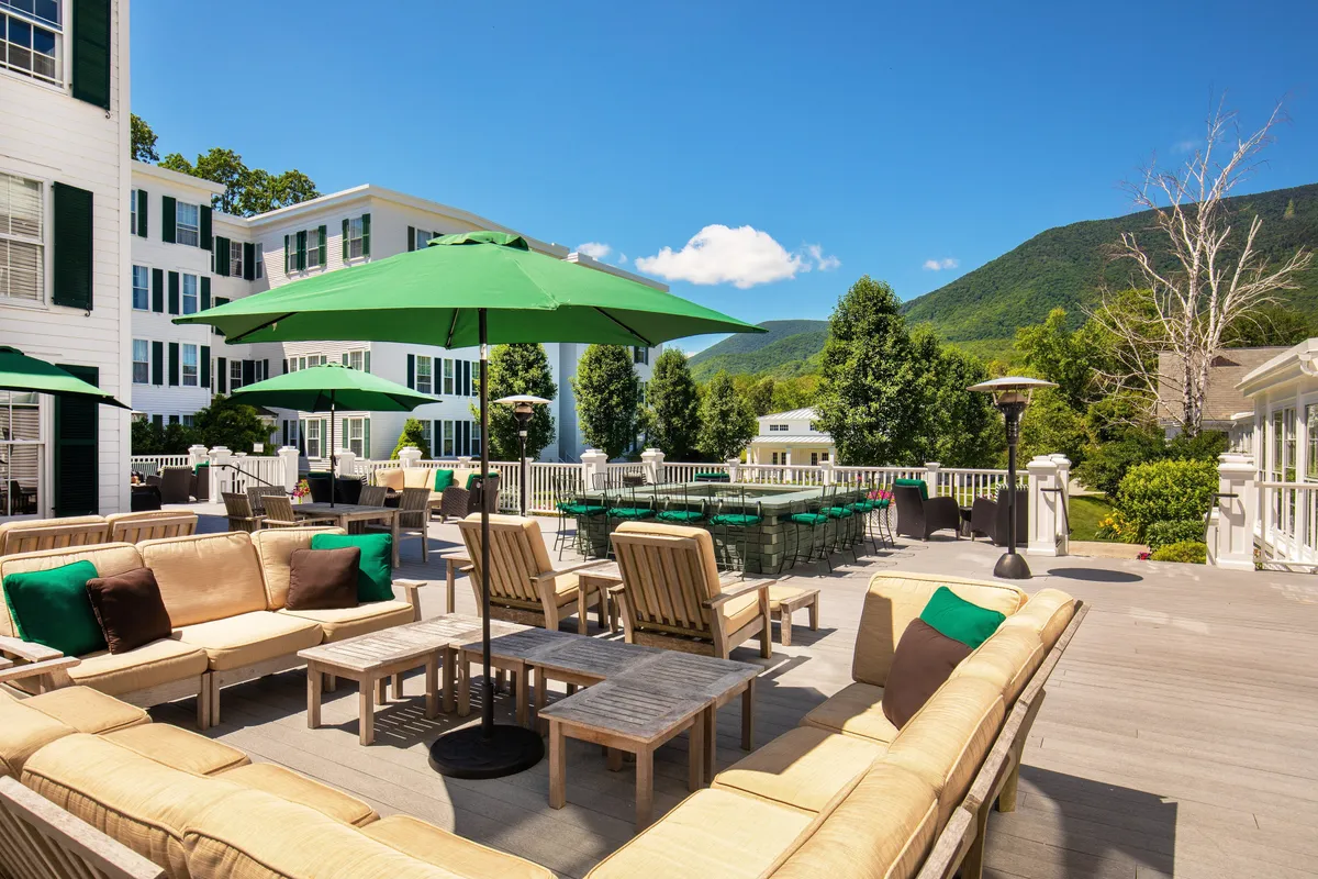 An inviting outdoor dining and lounge space with green umbrellas, ample seating, and picturesque mountain views. The area is set for casual dining and relaxing, with a clear sense of connection to nature and a backdrop that shifts with the seasons.