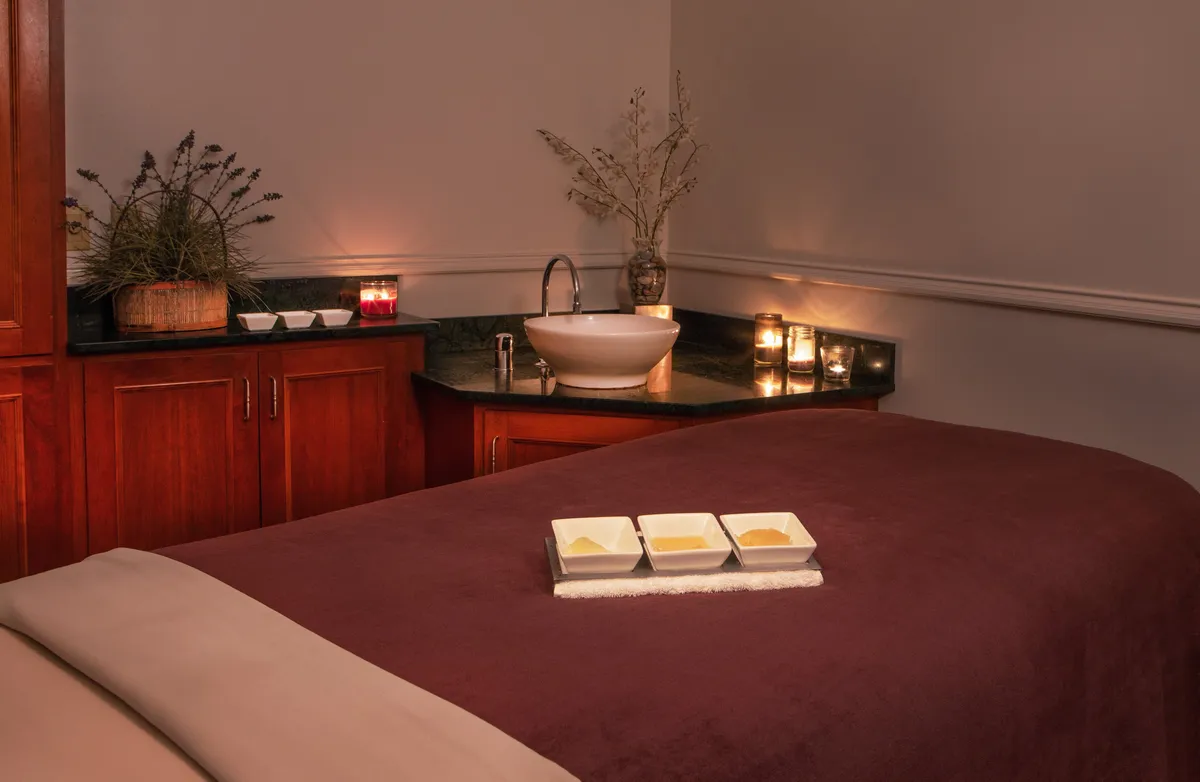 The scene shows a tranquil interior spa setting with a massage table, a bowl of oils, candles, and a sink. The lighting is soft and calming, reinforcing the atmosphere of relaxation and rejuvenation.