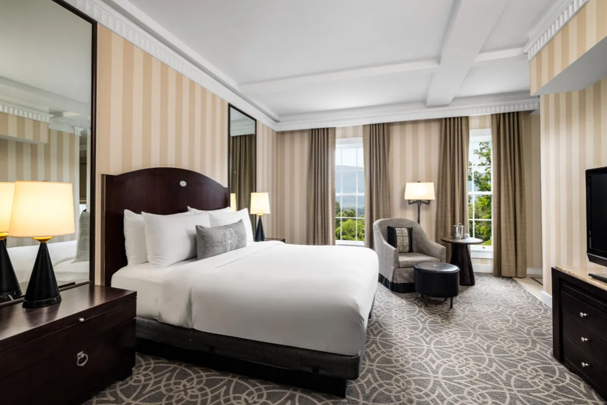 The image showcases a serene, light-filled hotel room with a plush king-sized bed as the centerpiece, flanked by a cozy accent chair, a stylish nightstand with a glowing lamp, and expansive windows that invite in natural light and offer scenic views.