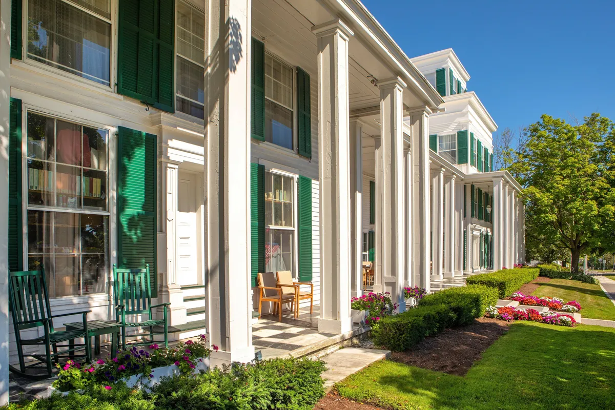 The image shows the inviting front porch of the resort, featuring classic green shutters, white columns, and an array of rocking chairs nestled among colorful flower beds. The scene exudes a sense of historic New England charm and offers a relaxing atmosphere perfect for unwinding or intimate conversations.