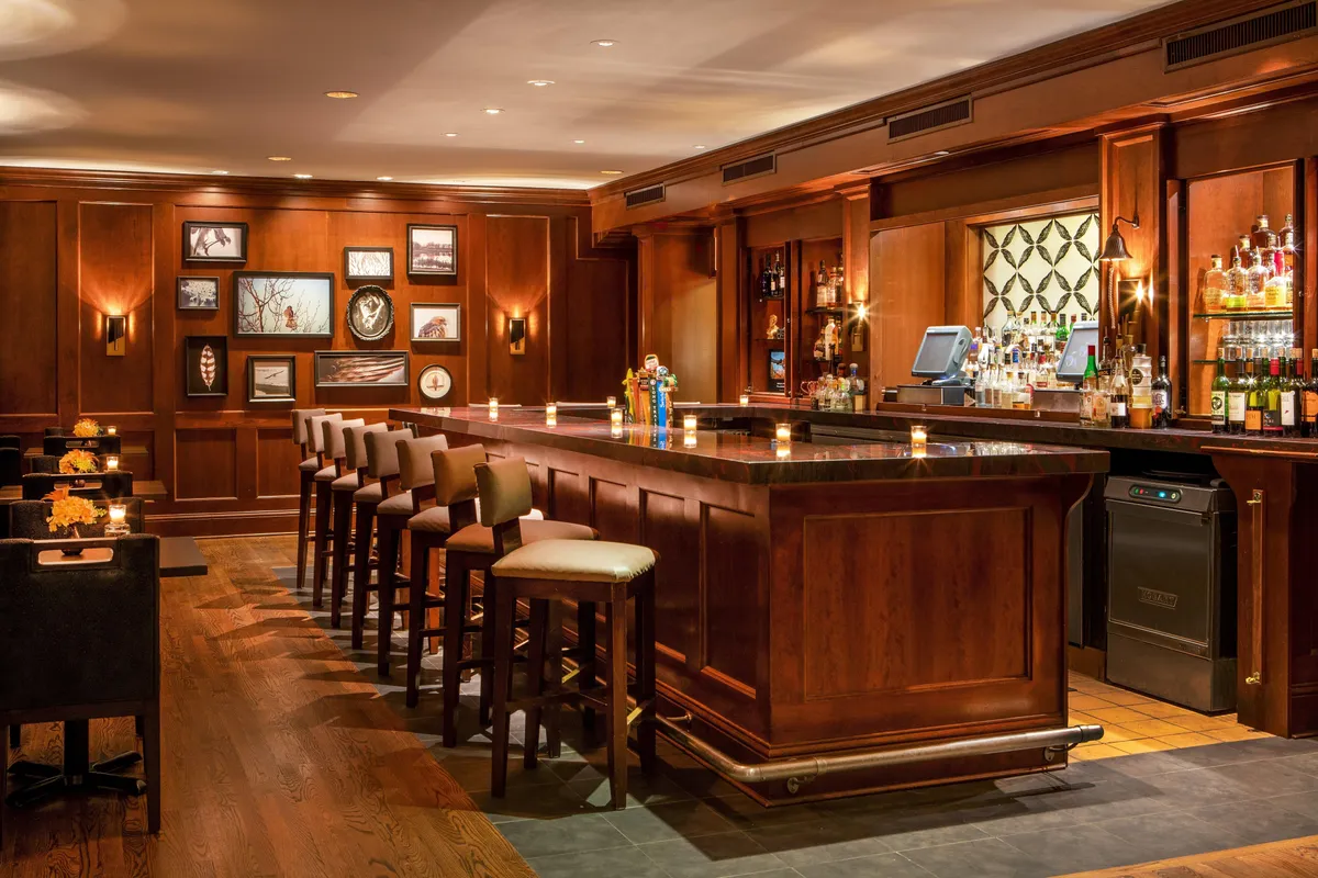 This interior shot captures a polished wooden bar lined with comfortable bar stools. Bottles are artfully displayed behind the bar, and framed artwork adds refinement to the ambiance. The space appears lively and welcoming, ideal for socializing, enjoying drinks, or relaxing after a day of outdoor activity.